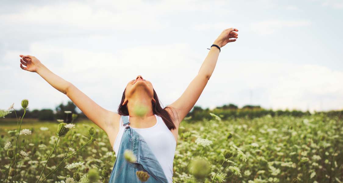A woman stands with her arms stretched up to the sky, looking up with a joyful expression in a wildflower meadow.