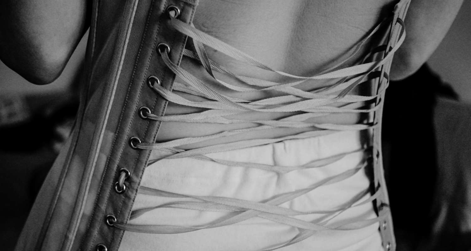 Detailed black and white image showing the back of a vintage corset, with its silk ribbon lacing hanging untied and ready to be tightened.