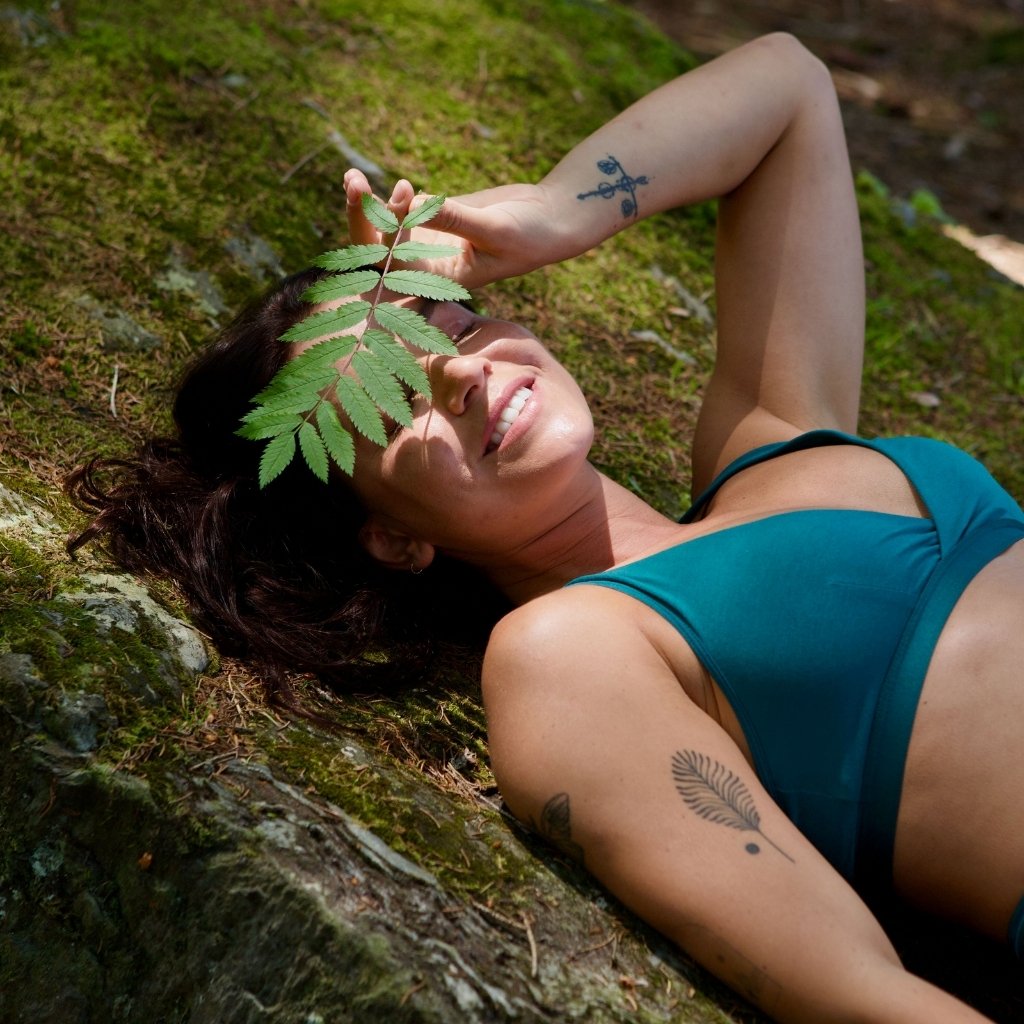 Model lying on a mossy rock in the forest wearing the Freya style in bayberry while playfully holding a leaf across her eyes, introducing our new limited edition colour.