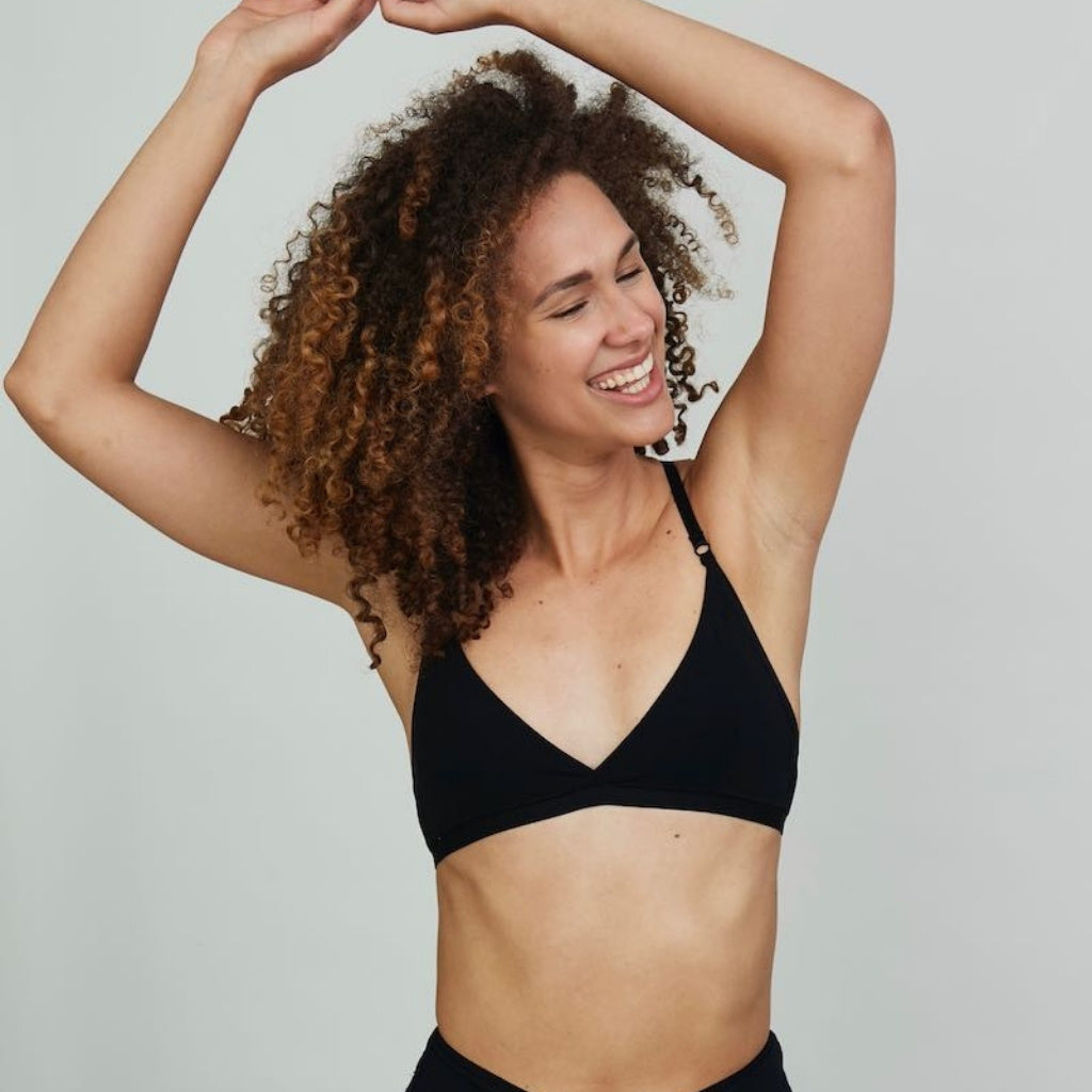 Model posing and smiling with her arms above her head wearing the Aurora bralette in black, showing one of our bestselling bralettes.